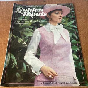 May include: A retro magazine cover for "Golden Hands" showing a woman in a pink outfit and hat. The cover advertises dressmaking and lampshade making. The magazine is displayed on a wooden surface.