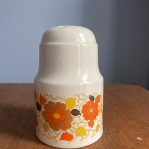 May include: A white ceramic salt shaker featuring a floral pattern in orange, yellow, and brown. The flowers and leaves are set against a cream background. The shaker has a rounded top with a single hole.