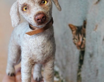 May include: A handmade felt dog figurine with a brown collar. The dog has light gray fur, a brown nose, and yellow eyes. A cat peeks out from behind a wall in the background. The dog is standing on a hand.
