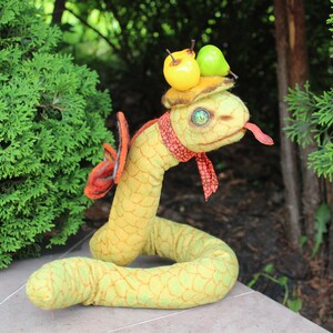 Op de afbeelding: Een handgemaakte pluche slang in geel en groen, met een patroon. Het heeft een rode sjaal, een hoed versierd met gele en groene vruchten en een rode tong. De slang heeft een groen oog.