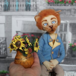 Può includere: Una bambola leone fatta a mano con occhi blu e una giacca blu. La bambola tiene in mano un piccolo vaso arancione pieno di girasoli gialli. Lo sfondo è un'immagine in bianco e nero di una scena di strada europea.
