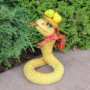 Può includere: Una statuetta di serpente in tessuto giallo con un motivo a scaglie testurizzato, che indossa un piccolo cappello beige sormontato da due pere in miniatura. Il serpente ha una lingua biforcuta rossa, occhi verdi e un nastro rosso annodato al collo.