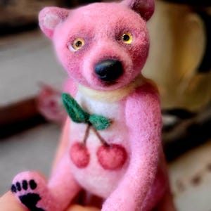 以下が含まれることがあります： 手作りのピンクのテディベア、チェリーのデザイン。クマは黒い鼻、黄色の目、小さな黄色の襟をしています。緑の葉が付いた2つの赤いさくらんぼがクマの胸に付いています。クマの足は黒です。