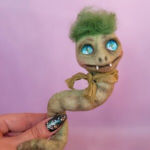 Può includere: Una creatura in feltro fatta a mano con un ciuffo verde, grandi occhi blu e una sciarpa beige. La creatura ha un corpo verde chiaro e due piccole zanne. Lo sfondo è viola chiaro.