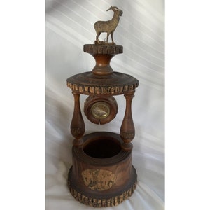 May include: A wooden clock with a carved deer on top and a clock face in the center. The clock is on a wooden base with a planter on the bottom.