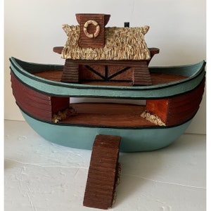May include: A wooden toy boat with a thatched roof, a life preserver, and a ramp. The boat is painted in shades of brown, red, and blue.