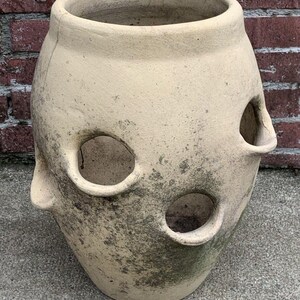 May include: A beige, weathered terracotta strawberry planter with a wide circular opening at the top and multiple smaller circular openings along its sides. The surface of the vessel shows signs of age, including dark, mottled discoloration.