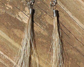 Op de afbeelding: Een paar oorhangers met lange, bruine haartufjes. De oorhangers zijn gemaakt van zilverkleurig metaal.