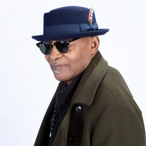 Puede incluir: Un hombre con un abrigo verde oscuro y un sombrero fedora azul marino con un adorno de pluma. Lleva gafas de sol y tiene una expresión seria.