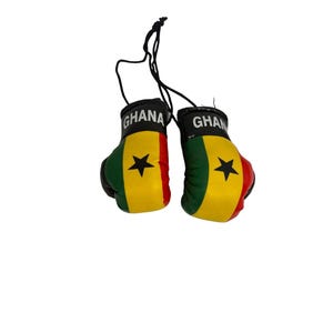 Peut inclure: Paire de mini gants de boxe aux couleurs du drapeau du Ghana : noir, vert, jaune et rouge. Chaque gant porte une étoile noire et le mot "GHANA" imprimé sur le poignet. Les gants sont reliés par une ficelle noire.