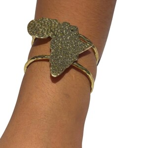 Puede incluir: Brazalete dorado con un mapa texturizado de África. El brazalete tiene un acabado de metal martillado y un diseño de tres bandas. El mapa es el punto focal, agregando un elemento cultural y geográfico a la joyería.