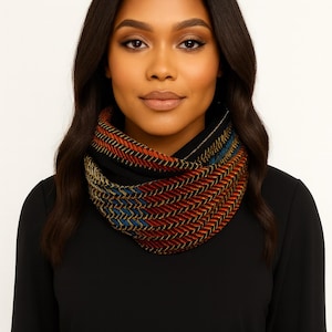 May include: A woman wearing a black cowl neck scarf with a colorful, patterned design. The scarf features a chevron pattern in shades of red, orange, blue, and gold. The scarf is made of a soft, woven material.