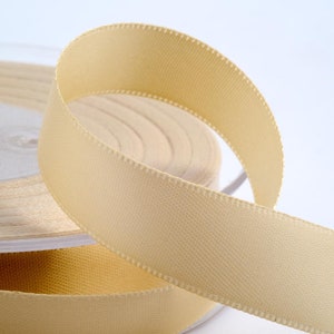 May include: Close-up of a spool of pale yellow satin ribbon. The ribbon is unwound, showing a smooth, slightly textured surface and a stitched edge. The spool is partially visible in the background, suggesting a continuous roll of ribbon.