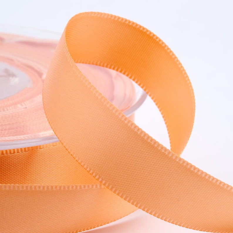 May include: A close-up shot of a spool of peach-coloured satin ribbon. The ribbon is unwound, showing a looped section with a stitched edge. The spool is visible in the background, suggesting a continuous roll of the ribbon. Suitable for crafts and gift wrapping.