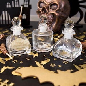 Könnte beinhalten: Drei durchsichtige Glasflaschen mit Korkstopfen und Etiketten auf einer schwarzen Oberfläche mit goldenem Glitzer und Halloween-Schnitten. Die Etiketten lauten "Potion d'Invisibilité".
