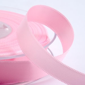 May include: A spool of pale pink satin ribbon. The ribbon is unravelling, revealing its smooth texture and stitched edges. The ribbon's soft pastel hue makes it suitable for crafts, gift wrapping, or decorative use.