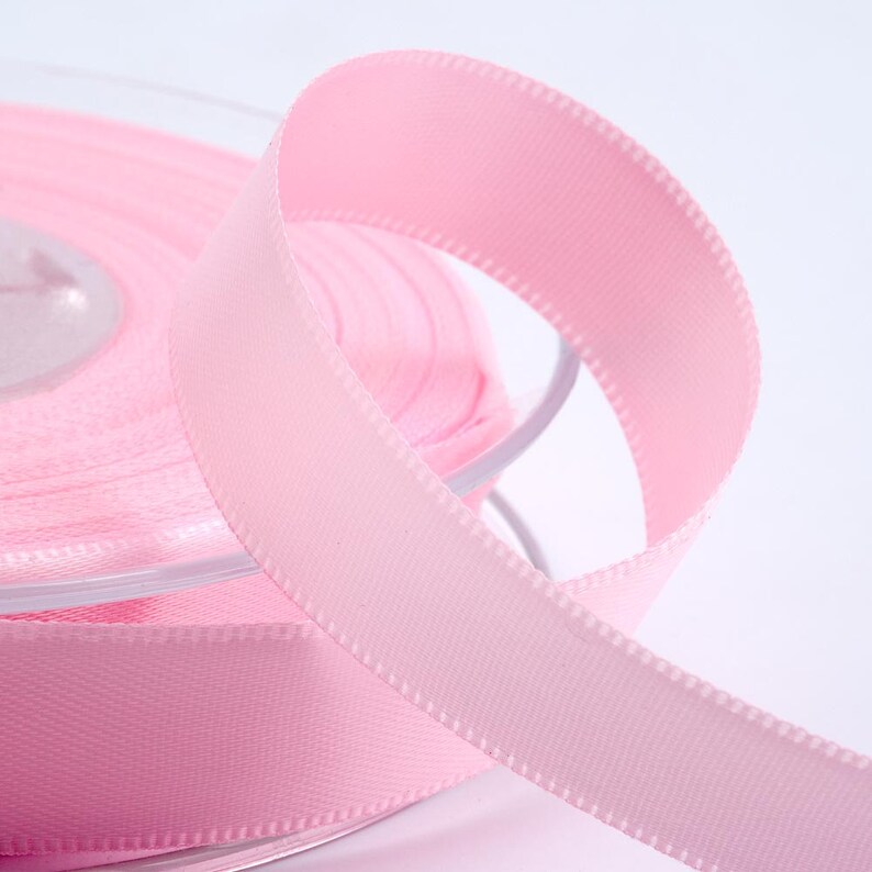 May include: A spool of pale pink satin ribbon. The ribbon is unravelling, revealing its smooth texture and stitched edges. The ribbon's soft pastel hue makes it suitable for crafts, gift wrapping, or decorative use.