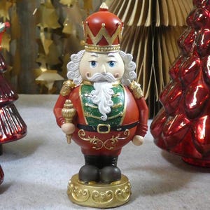 Könnte beinhalten: Dekorative Weihnachts-Nussknacker-Figur. Die Figur trägt einen roten Mantel mit goldenen Akzenten, eine grüne Weste und eine goldene Krone. Sie steht auf einem goldenen Sockel und hält ein Zepter. Rote Weihnachtsbaumornamente sind im Hintergrund.