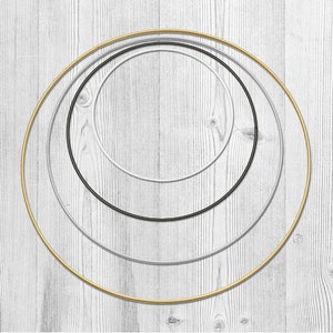 Könnte beinhalten: Vier Metallreifen in Gold, Silber, Schwarz und Weiß, konzentrisch auf einem hellen Holzmaserungshintergrund angeordnet. Die Reifen variieren in der Größe und erzeugen einen geschichteten Effekt. Ideal für Bastelarbeiten oder Dekorationszwecke.