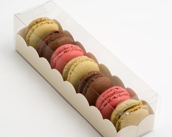 May include: A clear plastic box with a white scalloped border filled with colorful macarons. The macarons are arranged in a row and include pink, green, brown, and beige colors.