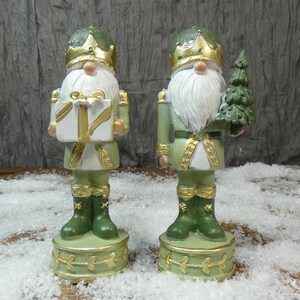 May include: Two Christmas nutcracker figures. One holds a gift box with gold ribbon, the other a small green tree. Both wear green and gold outfits, crowns, and have white beards. They stand on decorative bases.