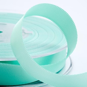 May include: A spool of pale turquoise ribbon. The ribbon is made of a smooth, woven material and is neatly coiled. The edges of the ribbon are subtly stitched. The ribbon is on a clear plastic spool, and the background is white.