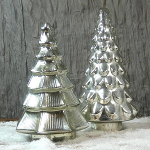 May include: Two silver Christmas tree ornaments. The trees have a layered design and sit on a base. The background is a textured grey fabric, and the base of the trees are on a bed of white snow.