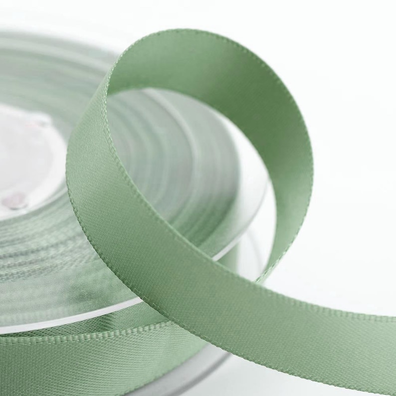 May include: A roll of sage green satin ribbon. The ribbon is tightly wound, with a section unspooled and curled, displaying a smooth, glossy texture. The ribbon's colour is a muted, earthy green, suitable for crafts and gift wrapping.