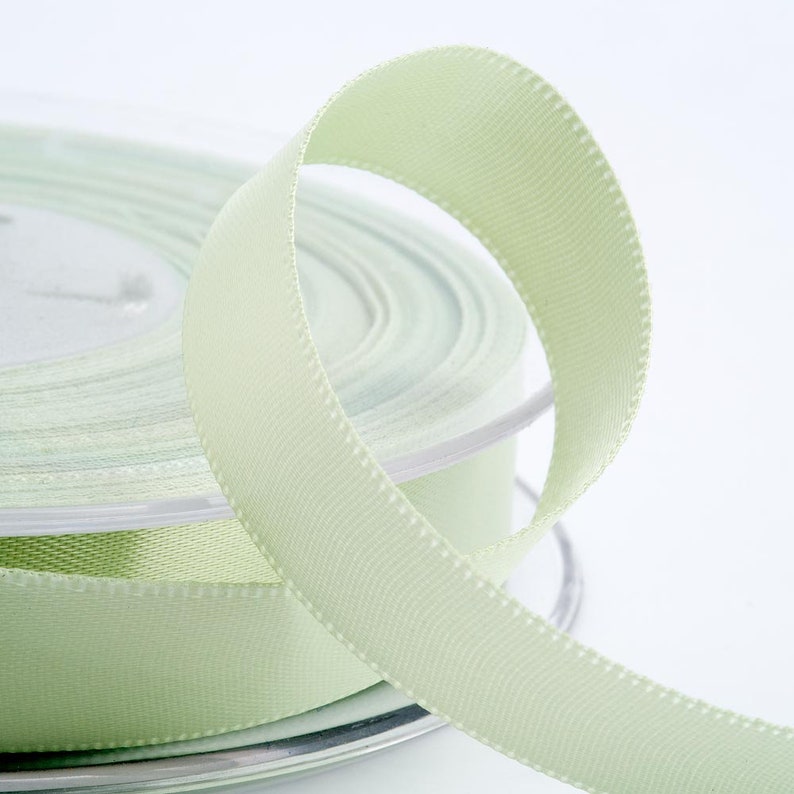 May include: A spool of pale green ribbon with a textured surface. The ribbon is unwound, forming a loop in the foreground. The spool is visible in the background, with the ribbon neatly coiled. The ribbon has a subtle sheen.