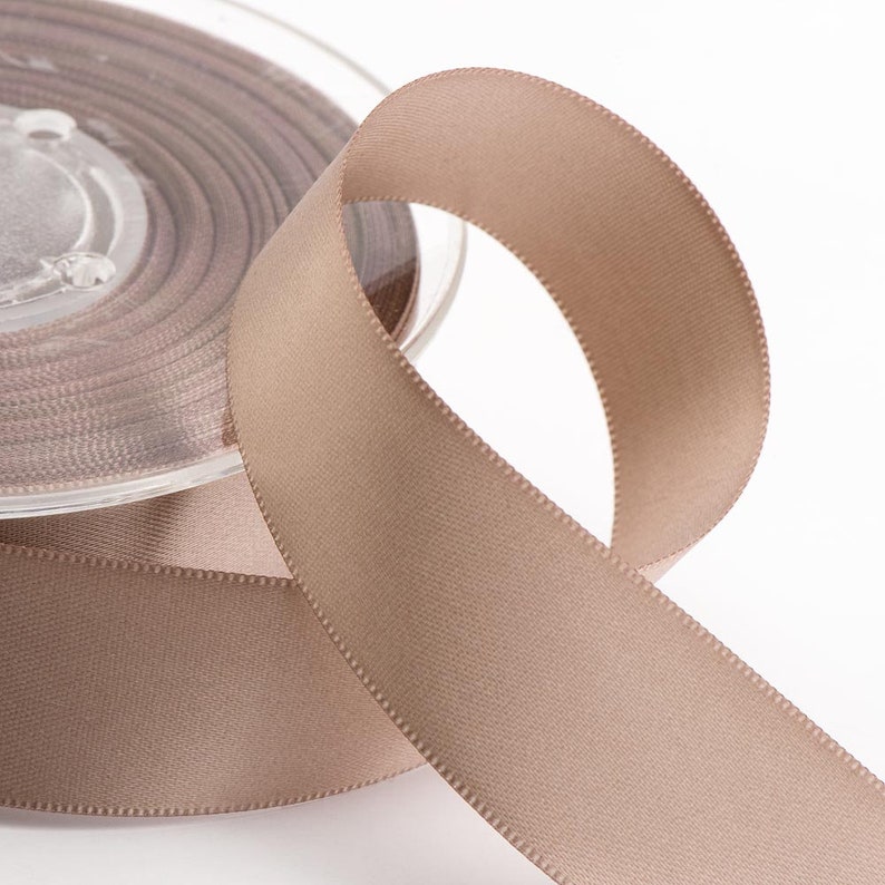 May include: A spool of taupe satin ribbon with a smooth, slightly shiny surface. The ribbon is unwound, revealing a loop against a white background. The spool is clear plastic, and the ribbon is tightly wound.