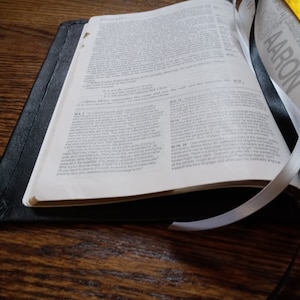 Puede incluir: Una Biblia encuadernada en cuero negro con marcadores de cinta blanca y amarilla. La Biblia est&aacute; abierta al libro de N&uacute;meros, cap&iacute;tulo 4, vers&iacute;culos 4-20. La palabra "AARON" es visible en la p&aacute;gina derecha.