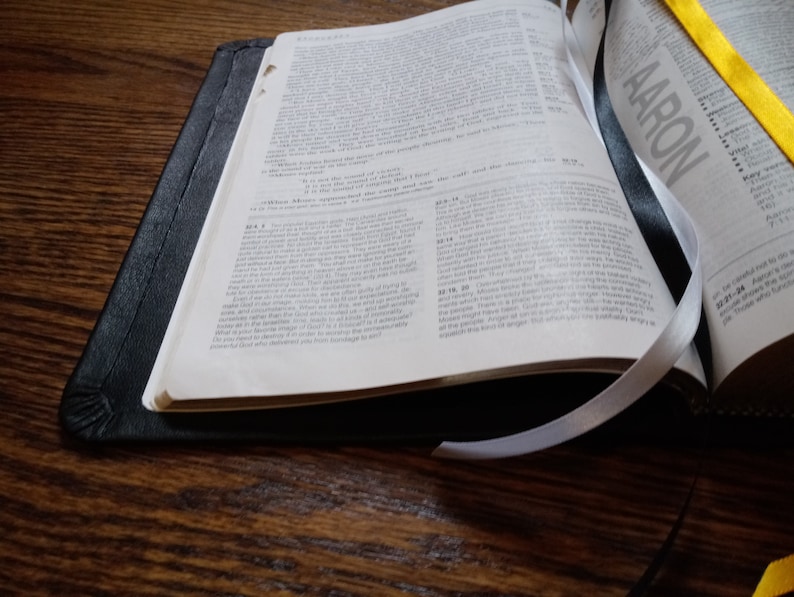 Puede incluir: Una Biblia encuadernada en cuero negro con marcadores de cinta blanca y amarilla. La Biblia est&aacute; abierta al libro de N&uacute;meros, cap&iacute;tulo 4, vers&iacute;culos 4-20. La palabra "AARON" es visible en la p&aacute;gina derecha.