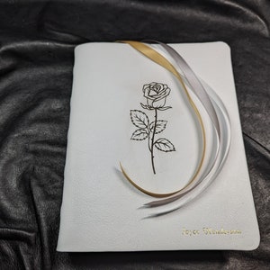 Puede incluir: Diario de cuero blanco con una cinta dorada y plateada atada alrededor. La portada presenta un dibujo de l&iacute;nea negra de una rosa y el texto "Joyce Henderson".