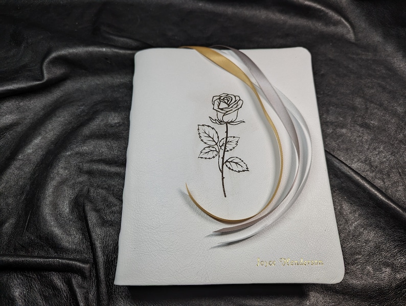 Puede incluir: Diario de cuero blanco con una cinta dorada y plateada atada alrededor. La portada presenta un dibujo de l&iacute;nea negra de una rosa y el texto "Joyce Henderson".