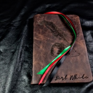 Puede incluir: Un diario de cuero marr&oacute;n con un dise&ntilde;o de pluma y tintero. El diario tiene una cinta negra, roja y verde atada alrededor. El nombre "Bert White" est&aacute; grabado en la parte delantera.