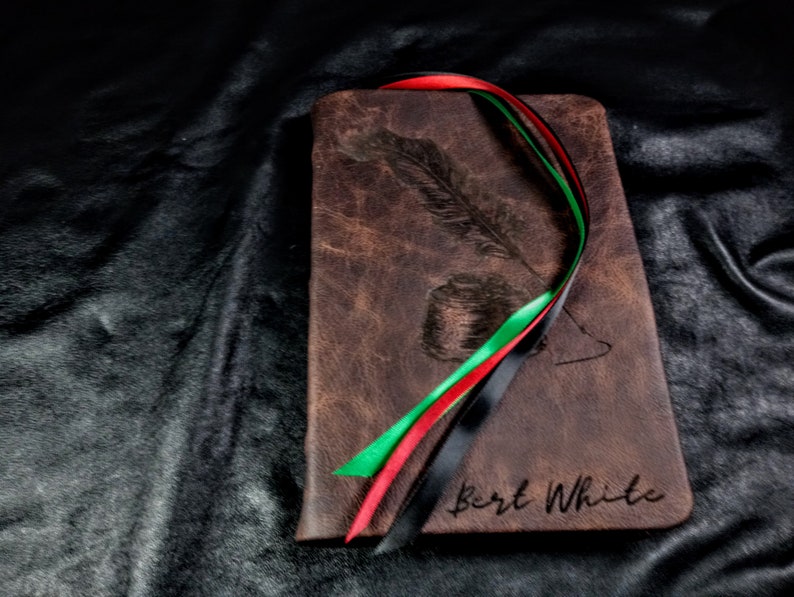 Puede incluir: Un diario de cuero marr&oacute;n con un dise&ntilde;o de pluma y tintero. El diario tiene una cinta negra, roja y verde atada alrededor. El nombre "Bert White" est&aacute; grabado en la parte delantera.