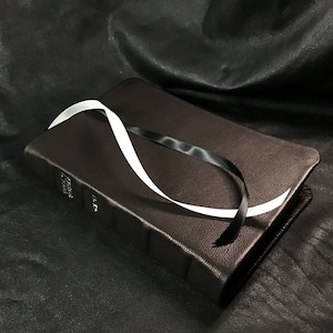 Puede incluir: Un libro encuadernado en cuero marr&oacute;n con un marcap&aacute;ginas de cinta blanca y negra. El libro est&aacute; sobre una superficie negra.