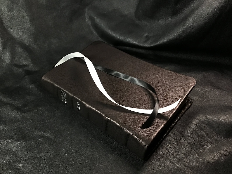 Puede incluir: Un libro encuadernado en cuero marr&oacute;n con un marcap&aacute;ginas de cinta blanca y negra. El libro est&aacute; sobre una superficie negra.