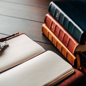 Könnte beinhalten: Ein offenes Notizbuch mit leeren Seiten, eine silberne Armbanduhr und ein Bleistift liegen auf einer dunklen Holzoberfläche. Drei ledergebundene Bücher in Marineblau, Rot und Hellbraun sind daneben gestapelt. Die Szene deutet auf einen Arbeitsbereich hin.
