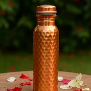 May include: A copper water bottle with a textured, dimpled surface. The bottle has a matching copper lid with a dark band. The bottle is on a wooden surface with scattered red and white flower petals. The background is blurred green.