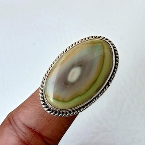 May include: A silver ring with an oval-shaped green, brown, and white agate gemstone. The ring has a rope-like design around the edge of the setting.