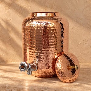May include: A cylindrical copper water dispenser featuring a hammered texture throughout the body and lid. The dispenser includes a metallic spigot with a black knob marked with the letters KK and a matching removable lid with a small central handle.
