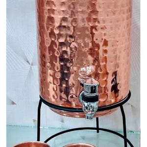 May include: Copper water dispenser with a hammered texture, a tap, and a lid. It stands on a black metal stand with two matching copper cups. The dispenser is designed for serving water or other beverages.
