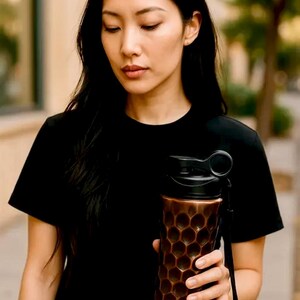 May include: A copper-coloured water bottle with a black lid and a honeycomb pattern. The bottle has a black strap and is held by a person wearing a black t-shirt. The background is blurred, suggesting an outdoor setting.
