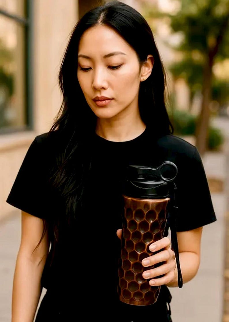 May include: A copper-coloured water bottle with a black lid and a honeycomb pattern. The bottle has a black strap and is held by a person wearing a black t-shirt. The background is blurred, suggesting an outdoor setting.