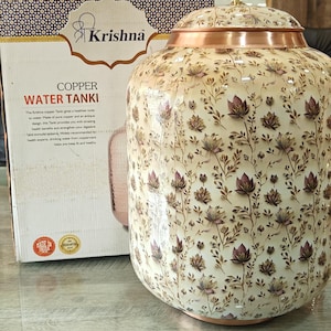 Könnte beinhalten: Ein Kupfer-Wassertank mit cremefarbener Außenseite und einem sich wiederholenden Blumenmuster in Braun- und Violetttönen. Der Tank hat einen abgerundeten Körper, einen kupferfarbenen Deckel mit kleinem Knauf und steht neben seiner Originalverpackung.