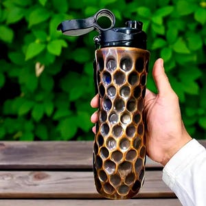 May include: A bronze-coloured water bottle with a honeycomb pattern and a black lid. The bottle is held in a hand against a backdrop of green foliage. The bottle has a flip-top opening.