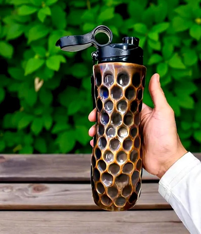 May include: A bronze-coloured water bottle with a honeycomb pattern and a black lid. The bottle is held in a hand against a backdrop of green foliage. The bottle has a flip-top opening.