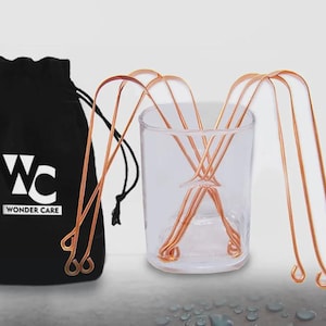 Puede incluir: Un juego de raspadores de lengua de color cobre en un vaso transparente y una bolsa negra con cordón. La bolsa tiene el texto blanco "WC" encima de "WONDER CARE". Los raspadores de lengua tienen un diseño curvo con asas en bucle.