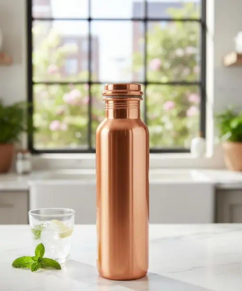 May include: A copper-colored water bottle with a cylindrical shape. The bottle is made of metal and has a screw-on cap. A glass of water with ice and mint sits next to the bottle on a white countertop.